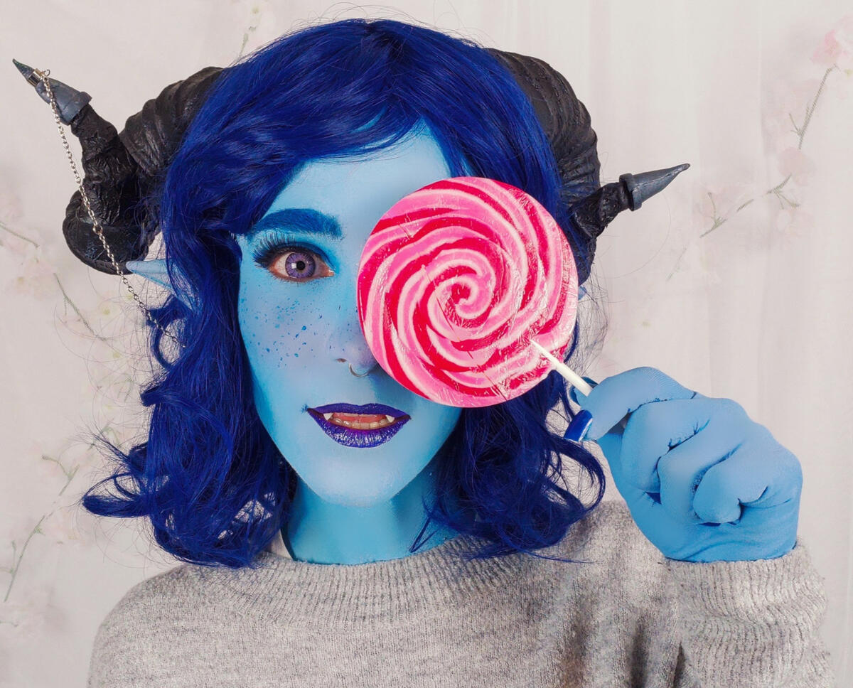 Photo of me dressed as Jester. I'm wearing a blue, shoulder length, wavy wig; black ram horns. My skin is painted blue.
